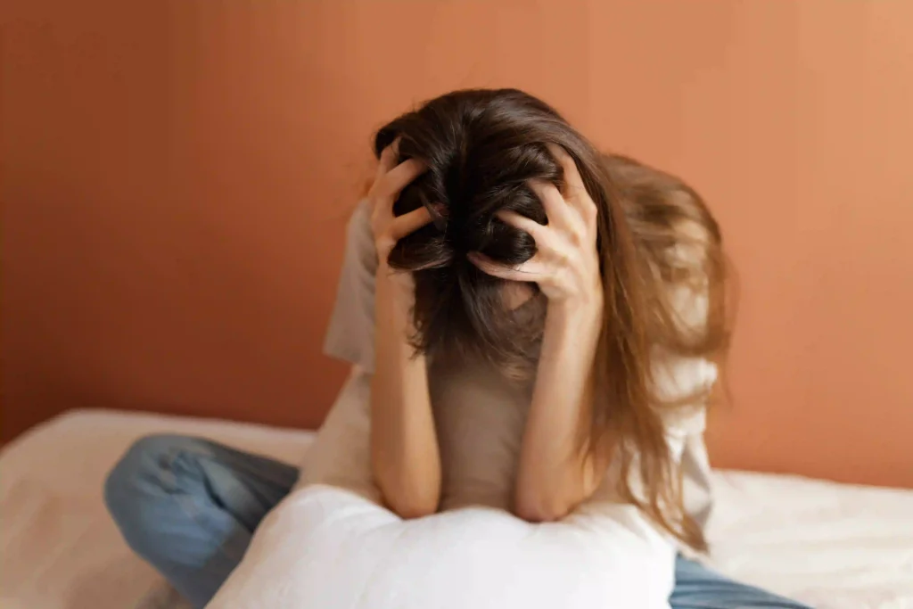 A woman is seated on a bed, her hands placed on her head, reflecting a moment of thought or concern.